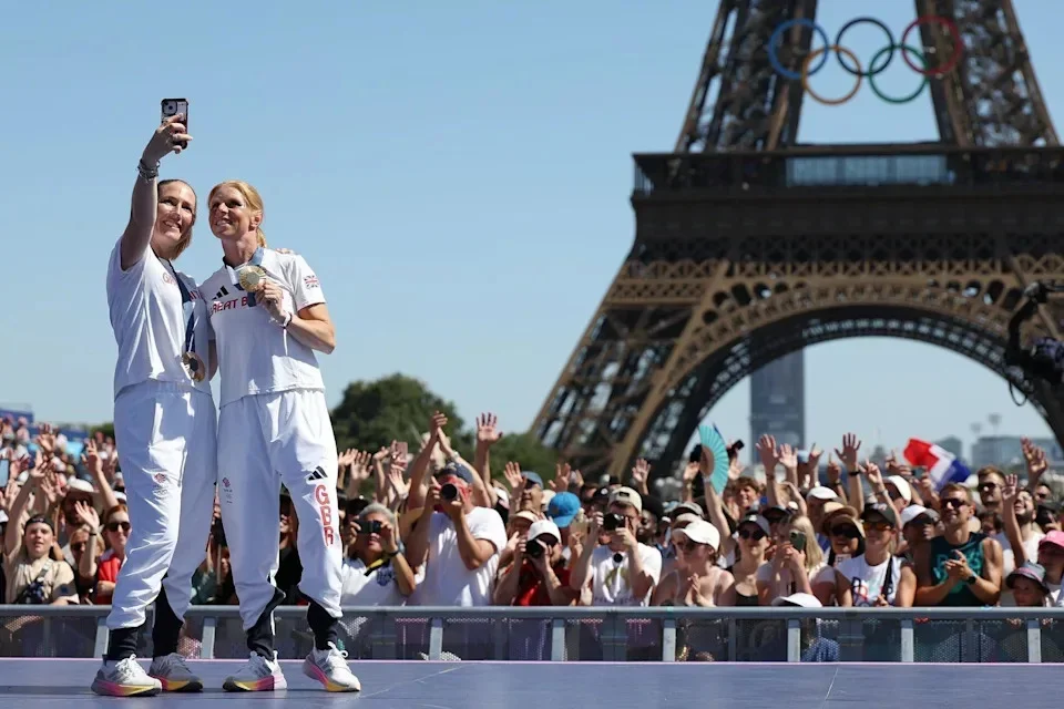 Equestrian eventing team gold medallists Ros Canter and Laura Collett pose for a photo at the Olympic Games in Paris in 2024. There are crowds of people in the background, standing in the shadow of the Eiffel Tower.