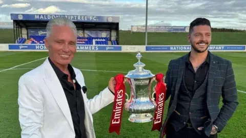 Warren Muggleton/BBC Barrie and Scott Drewitt-Barlow are holding the FA Cup - which is a silver trophy with red ribbons on it - on the pitch at Maldon and Tiptree FC. Behind them is a small blue stand. Barrie is wearing a black shirt and a white blazer. Scott is wearing a checkered dark grey suit.